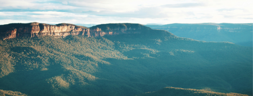 Sunlit escarpment above expansive forested valleys under a bright, partly cloudy sky.
